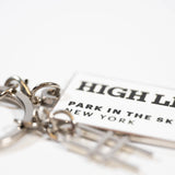 Closeup of the High Line Tag Keychain, featuring a small charm of the High Line railroad-track "H" icon out of focus, with the tag featuring the High Line wordmark and "Park in the Sky, New York NY" in black on white enamel in focus.