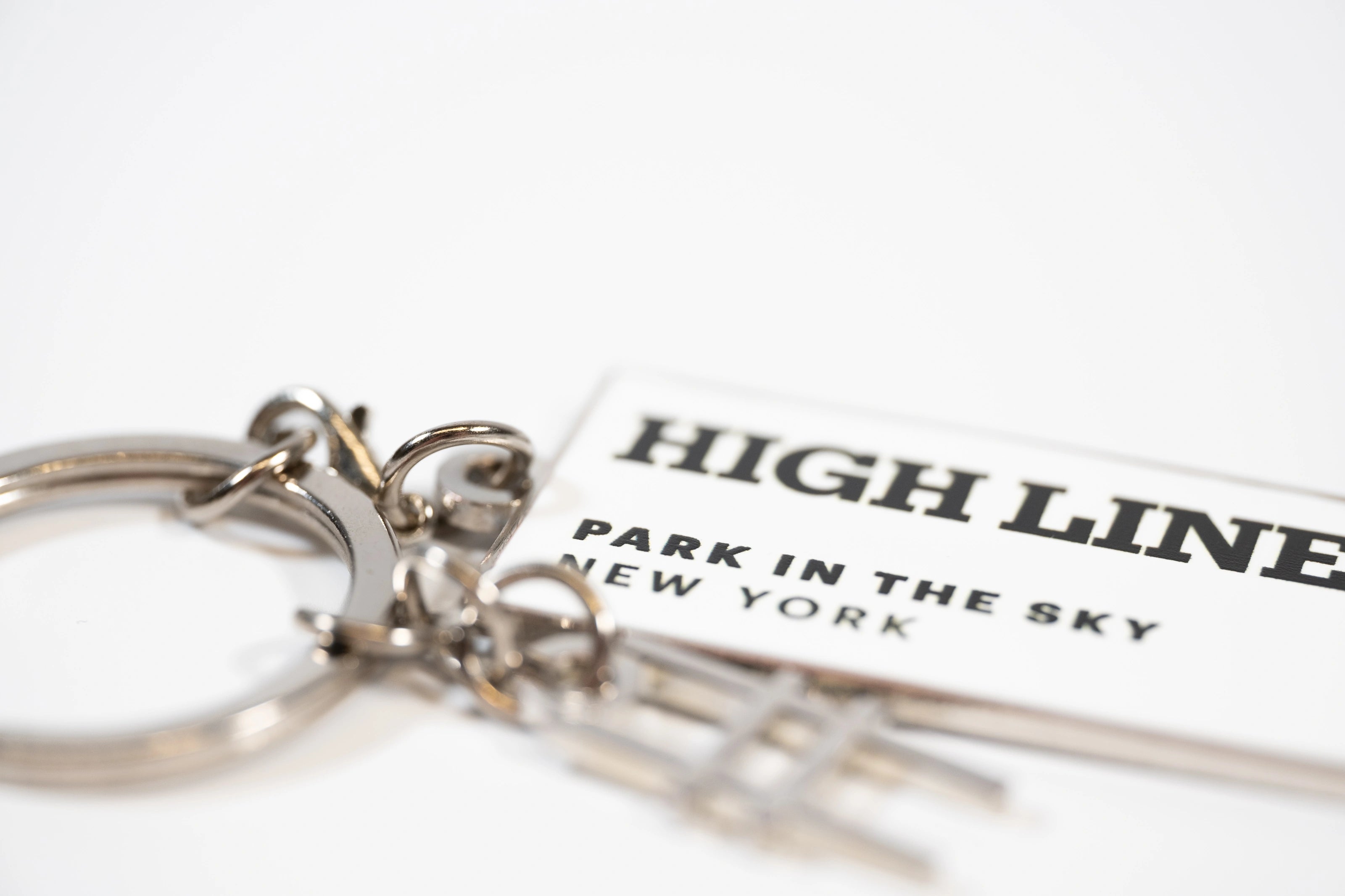 Closeup of the High Line Tag Keychain, featuring a small charm of the High Line railroad-track "H" icon out of focus, with the tag featuring the High Line wordmark and "Park in the Sky, New York NY" in black on white enamel in focus.