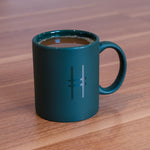 High Line Mug in Ivy, full of coffee, sitting on a wood desk. The mug is a deep green and matte with the High Line Icon visible in gloss on the side that's shown.