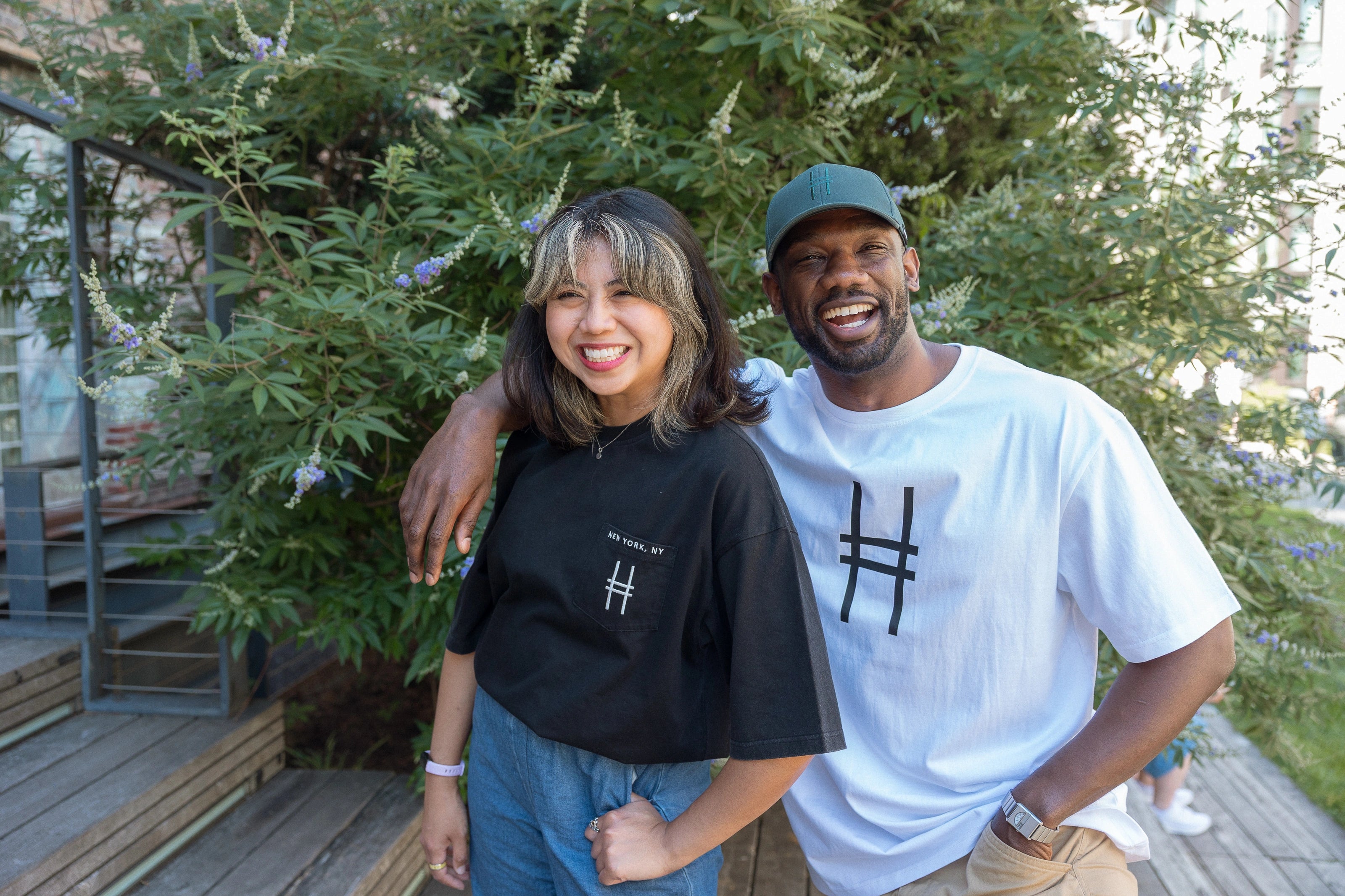 Two people smiling outdoors on the High Line at the 23rd street steps. The person on the right wears a the High Line Icon Tee in White and the High Line Icon Cap in Ivy, while the person on the left wears the Embroidered Icon Pocket Tee in Black.