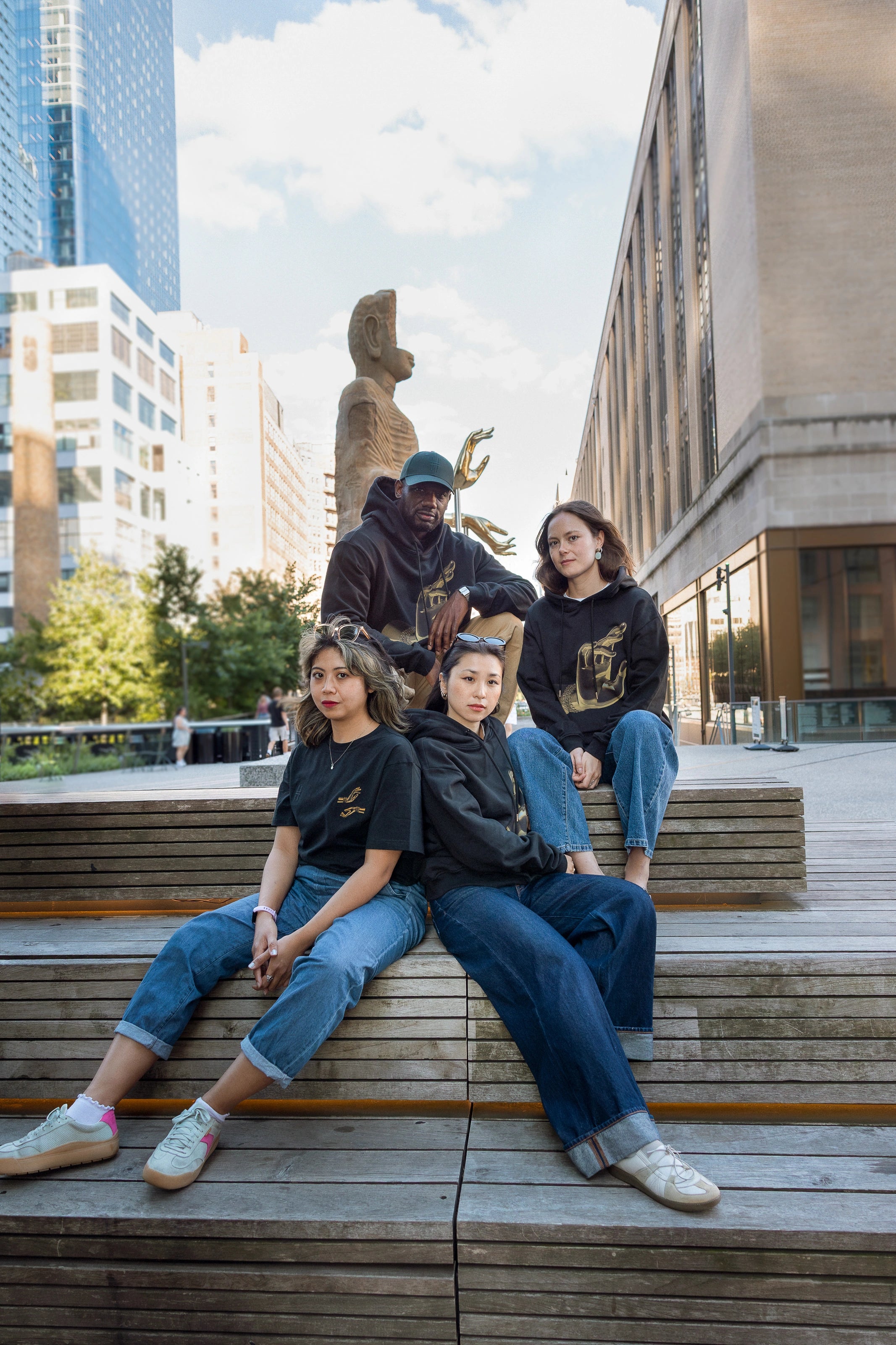 Four people, each in either the Light That Shines... Tee or Hoodie sit together in front of The Light That Shines Through the Universe statue by Tuan Andrew Nguyen on the High Line.