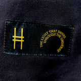 Closeup on the tag sewn onto the bottom left front of the hoodie—a black label with the High Line icon and "The Light That Shines Through the Universe" in gold.