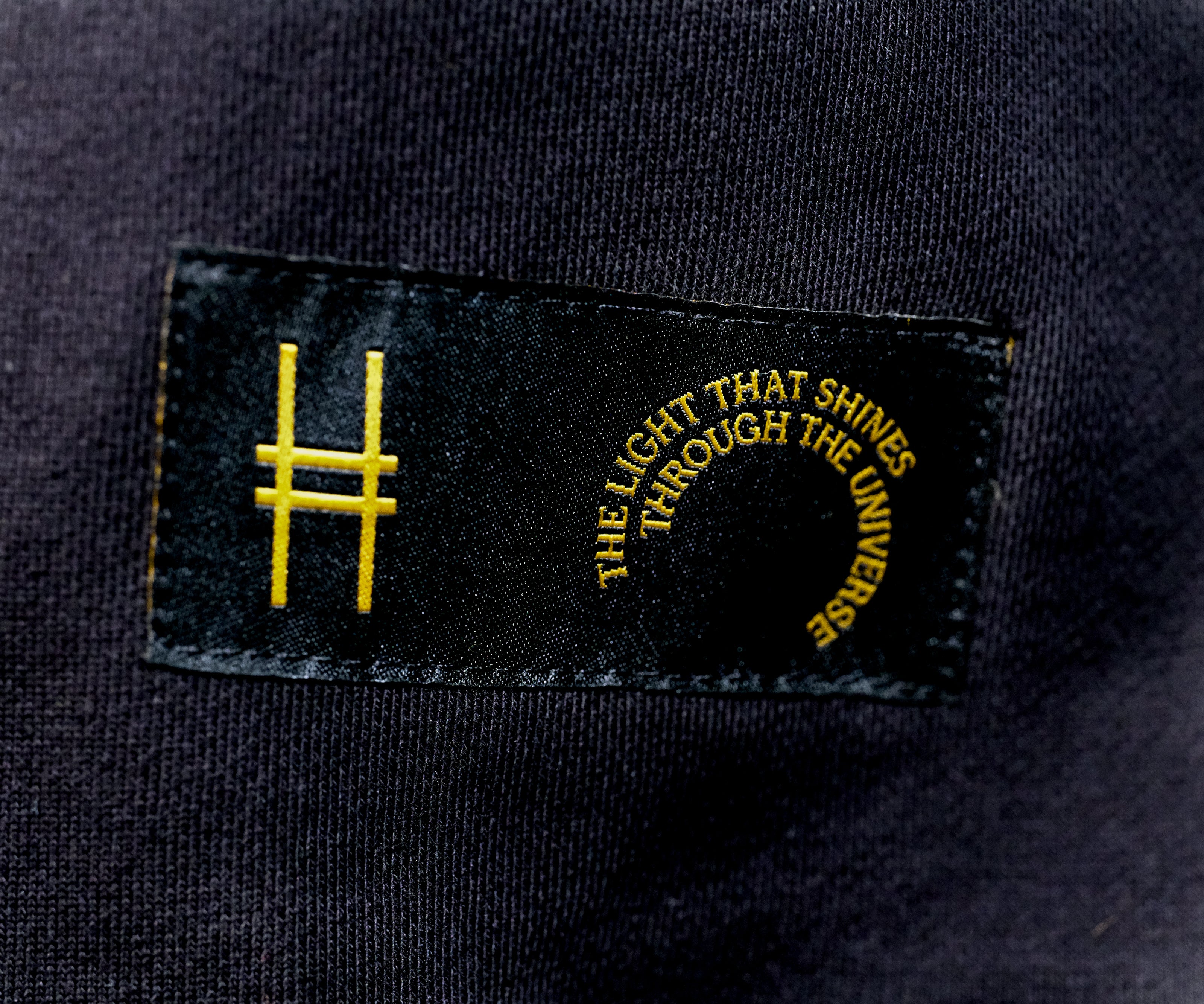 Closeup on the tag sewn onto the bottom left front of the hoodie—a black label with the High Line icon and "The Light That Shines Through the Universe" in gold.