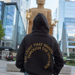 Person in The Light That Shines... Hoodie standing in front of the statue itself, with the back of the hoodie visible. The hoodie is emblazoned with the title of the piece in gold.