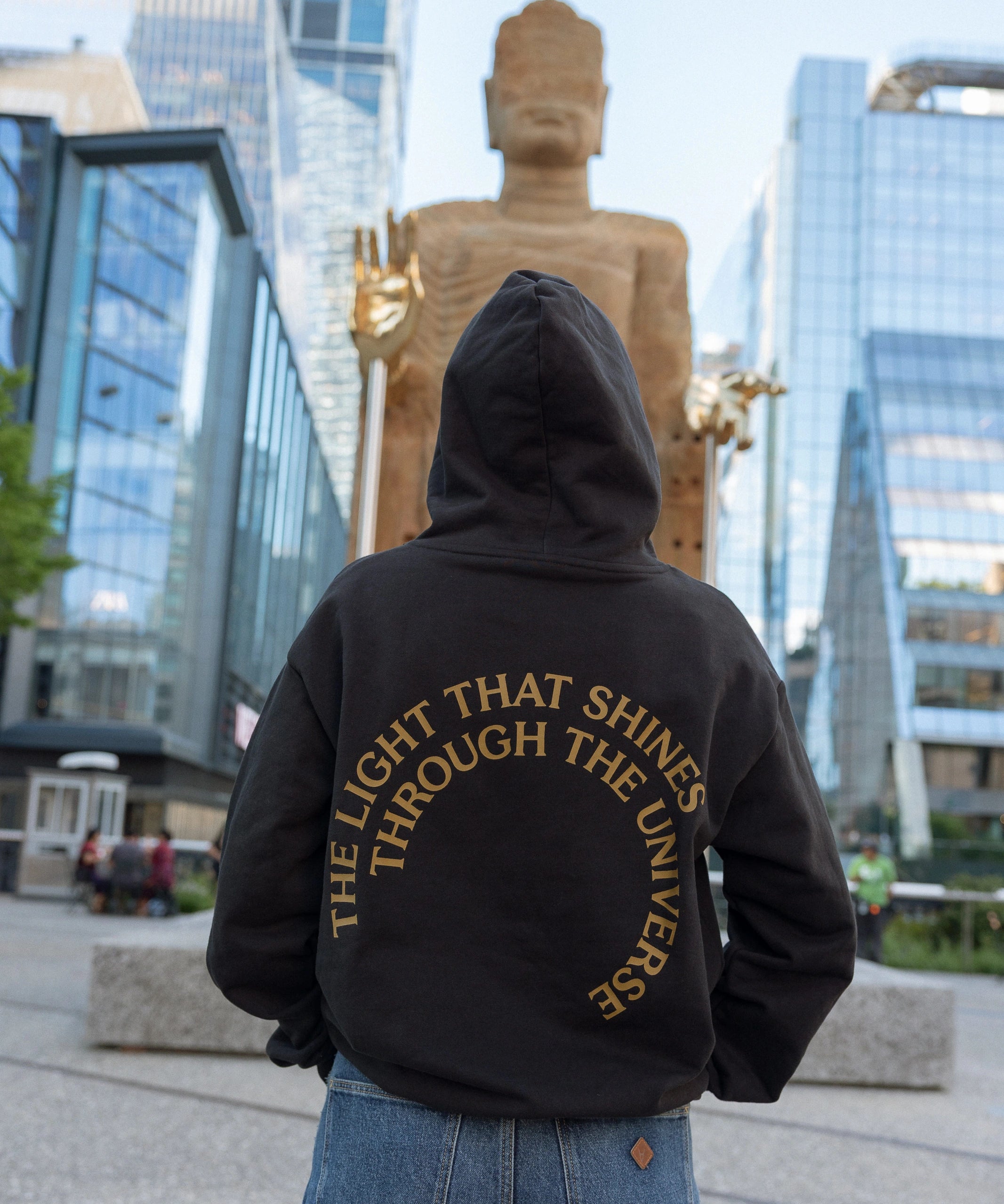 Person in The Light That Shines... Hoodie standing in front of the statue itself, with the back of the hoodie visible. The hoodie is emblazoned with the title of the piece in gold.