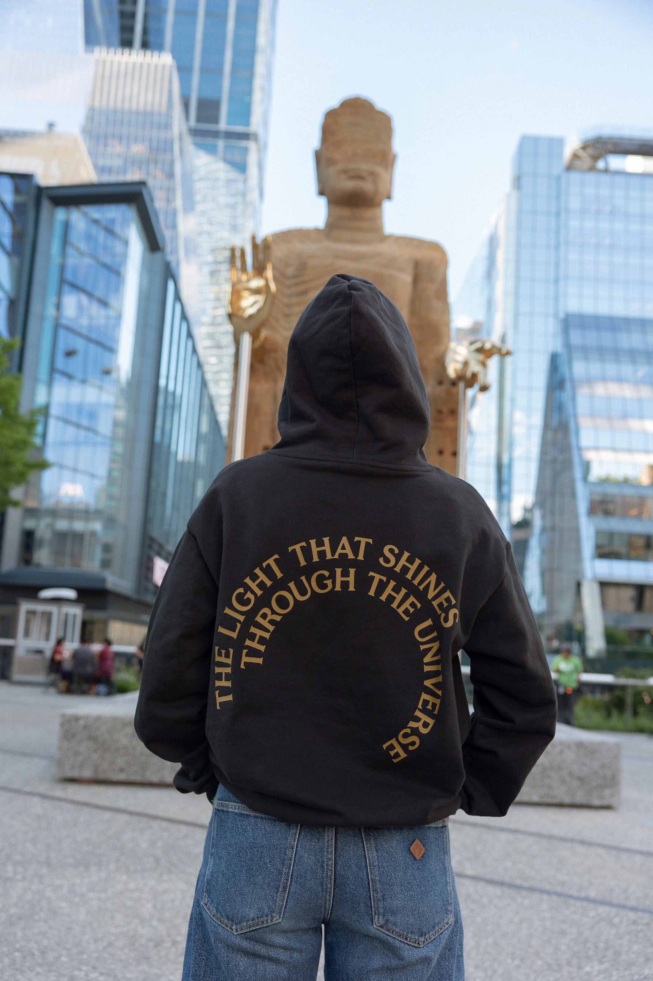 Person in The Light That Shines... Hoodie standing in front of the statue itself, with the back of the hoodie visible. The hoodie is emblazoned with the title of the piece in gold.