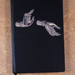 The Light That Shines... notebook. A black notebook featuring an image of statue's gold hands, each in a ritual position or mudra. Abhaya mudra signifies fearlessness and Varada mudra signifies compassion. 