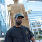 Man wearing The Light That Shines... Tee, standing in front of the statue itself. Both the statue's large brass hands and the embroidered hands on the t-shirt are visible.