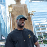Man wearing The Light That Shines... Tee, standing in front of the statue itself. Both the statue's large brass hands and the embroidered hands on the t-shirt are visible.