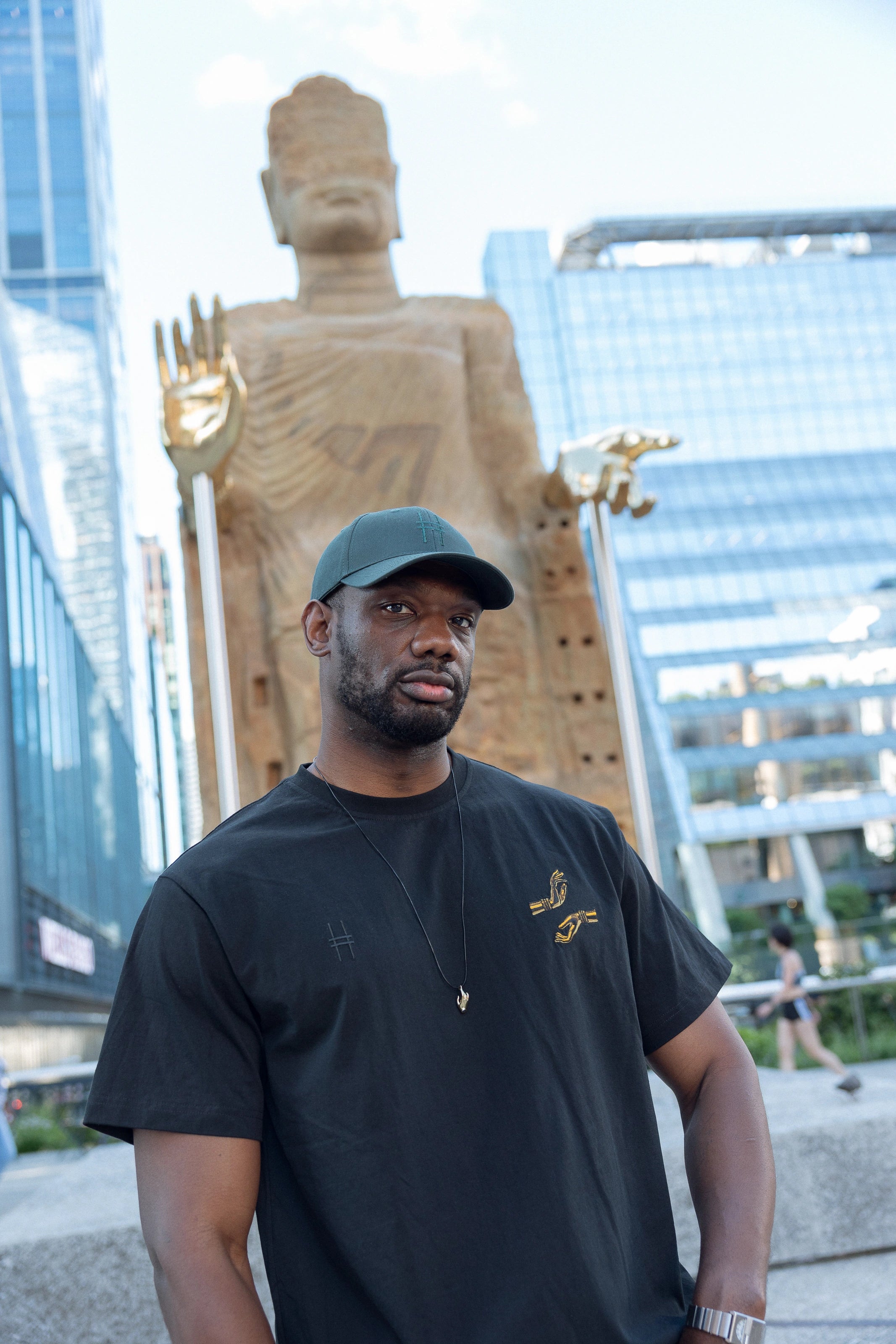 Man wearing The Light That Shines... Tee, standing in front of the statue itself. Both the statue's large brass hands and the embroidered hands on the t-shirt are visible.
