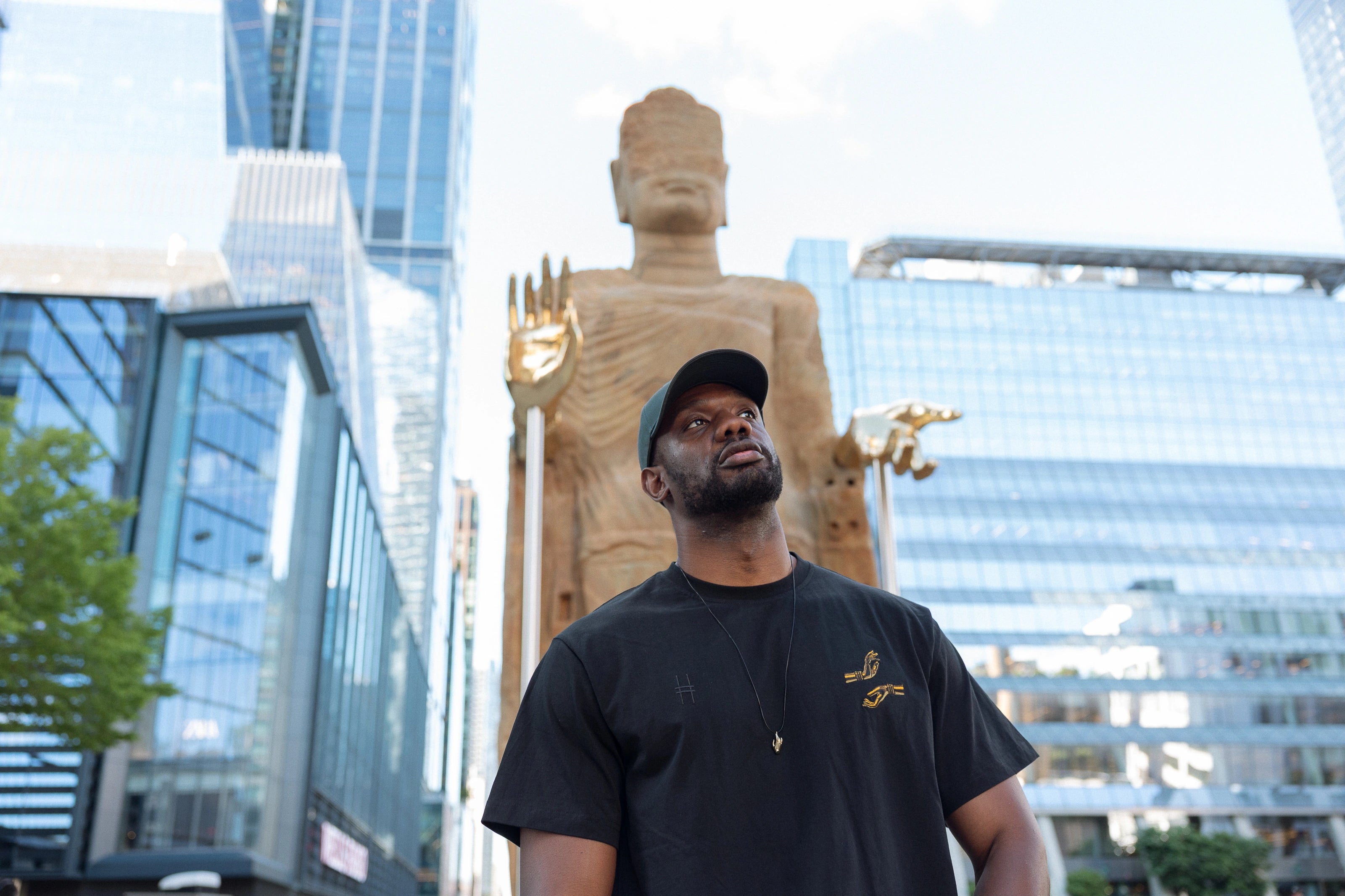 Man wearing The Light That Shines Tee, a black t-shirt featuring embroidery of the statues brass hands, standing in front of the statue on the High Line.