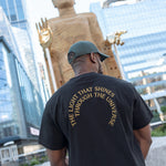 Man wearing The Light That Shines... Tee, facing the statue itself, so the title of the piece in gold across the back of the t-shirt is visible.
