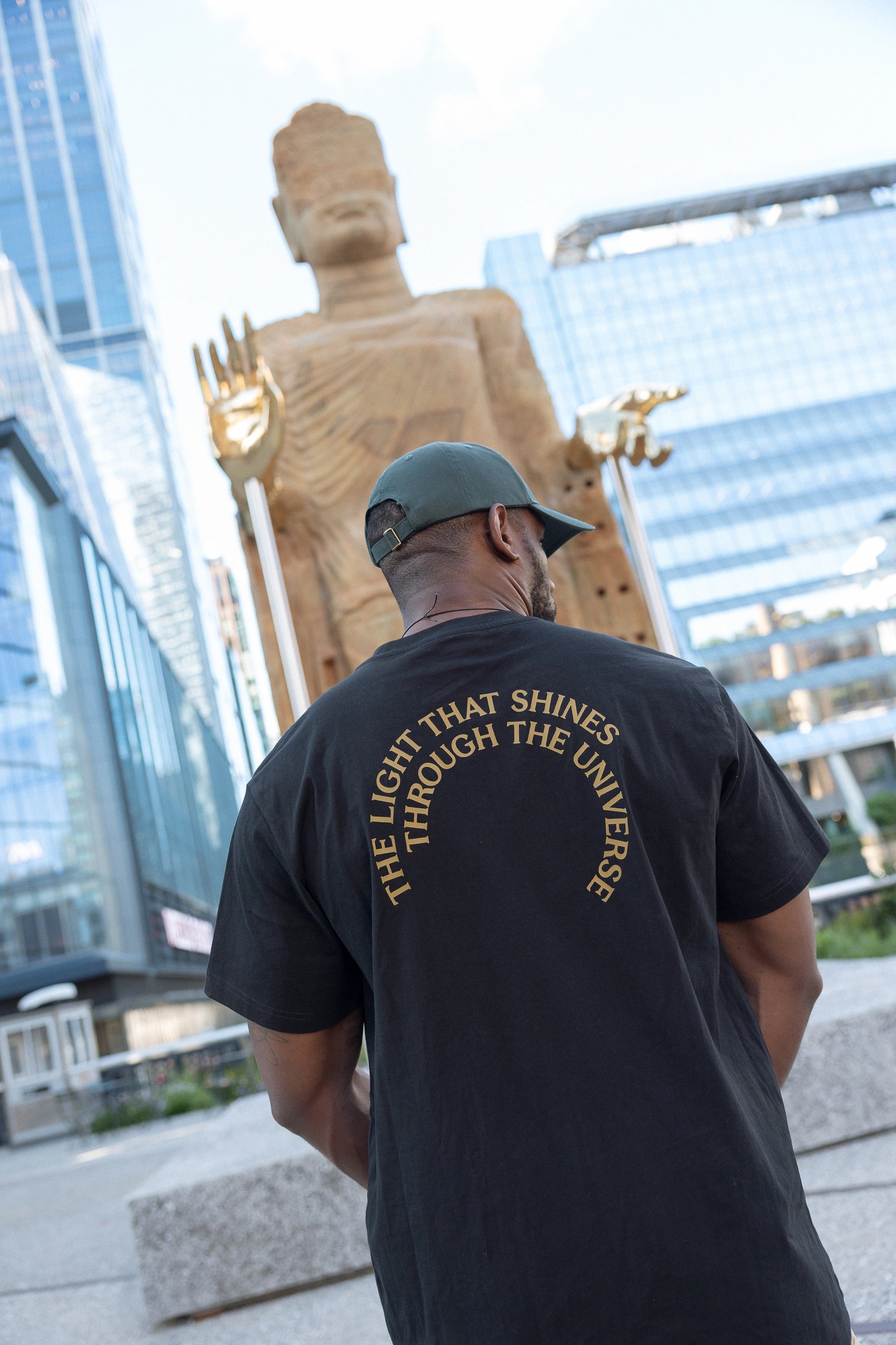 Man wearing The Light That Shines... Tee, facing the statue itself, so the title of the piece in gold across the back of the t-shirt is visible.
