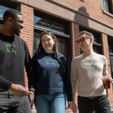 Three people walking from the Lingua Franca store on Washington & Jane. They each wear a Lingua Franca x High Line Sweater. These sweaters, two cotton-cashmere blends and one 100% cotton, are gender neutral and feature the phrase "park in the sky" embroidered in green cursive on the chest.