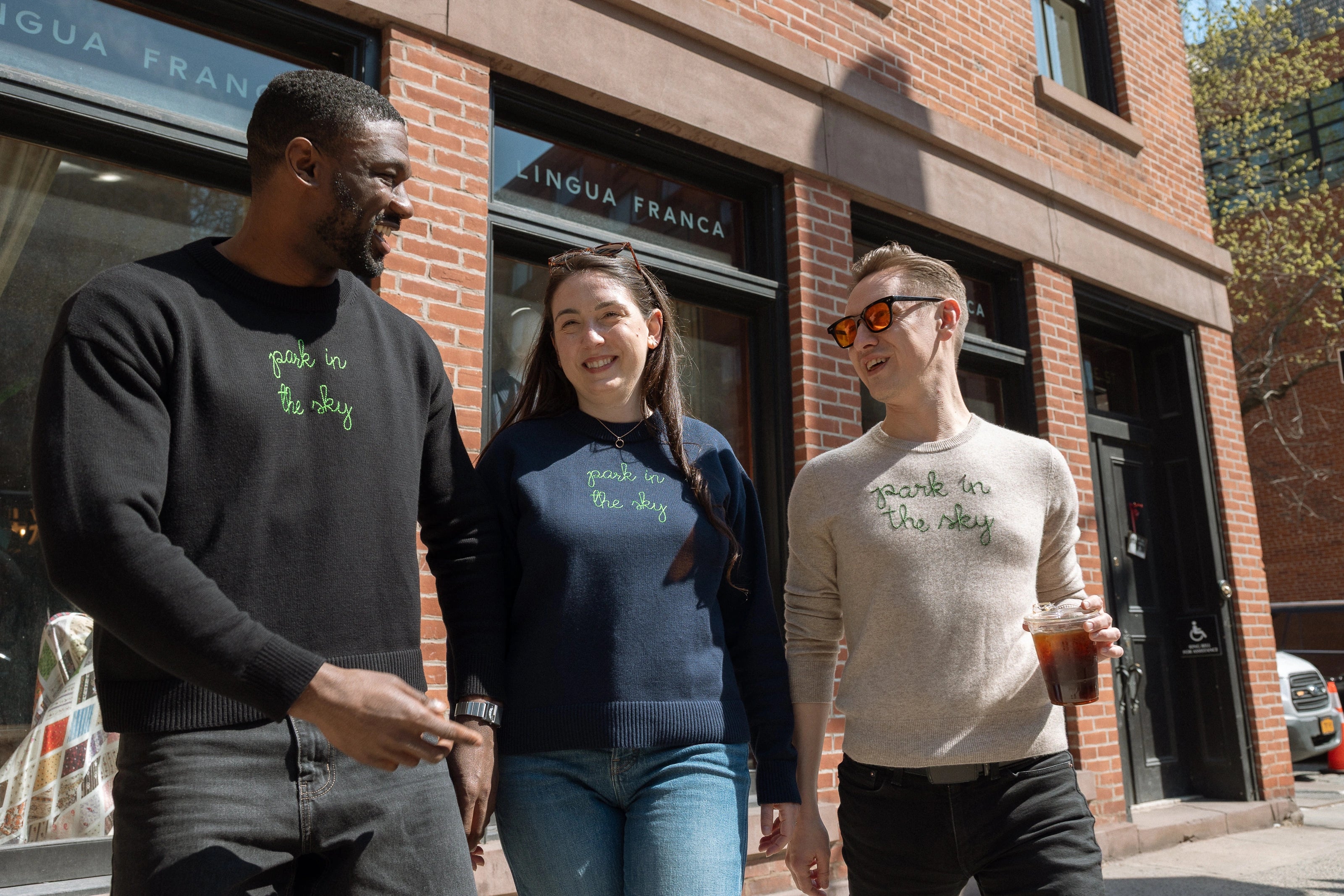 Three people walking from the Lingua Franca store on Washington & Jane. They each wear a Lingua Franca x High Line Sweater. These sweaters, two cotton-cashmere blends and one 100% cotton, are gender neutral and feature the phrase "park in the sky" embroidered in green cursive on the chest.
