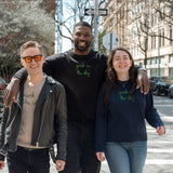 Three people walking from the High Line down Washington Street towards the Lingua Franca store. They each wear a Lingua Franca x High Line Sweater. These sweaters, two cotton-cashmere blends and one 100% cotton, are gender neutral and feature the phrase "park in the sky" embroidered in green cursive on the chest.
