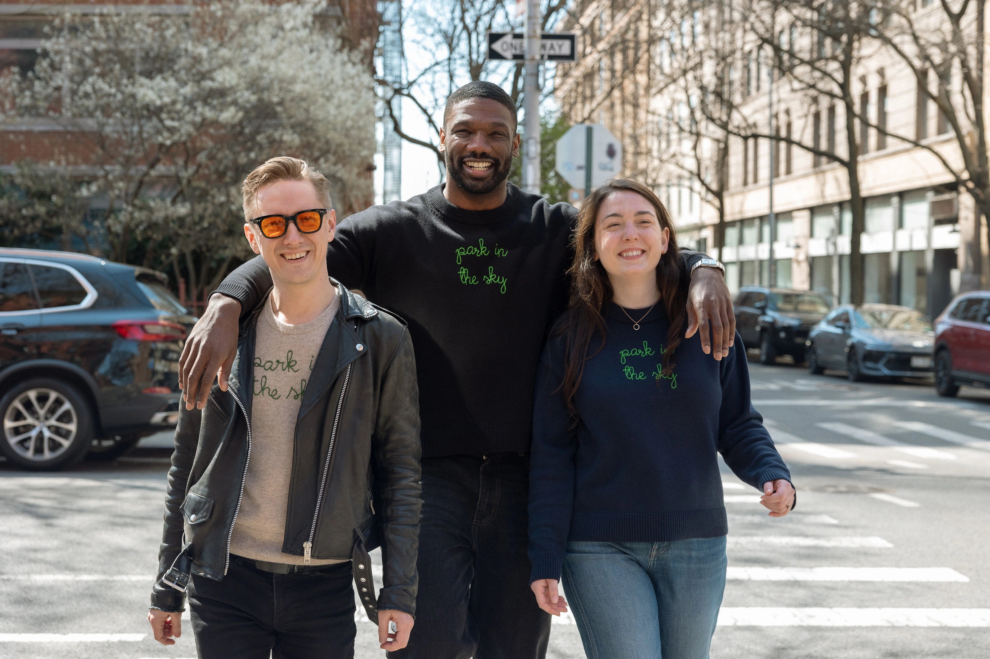 Three people walking from the High Line down Washington Street towards the Lingua Franca store. They each wear a Lingua Franca x High Line Sweater. These sweaters, two cotton-cashmere blends and one 100% cotton, are gender neutral and feature the phrase "park in the sky" embroidered in green cursive on the chest.