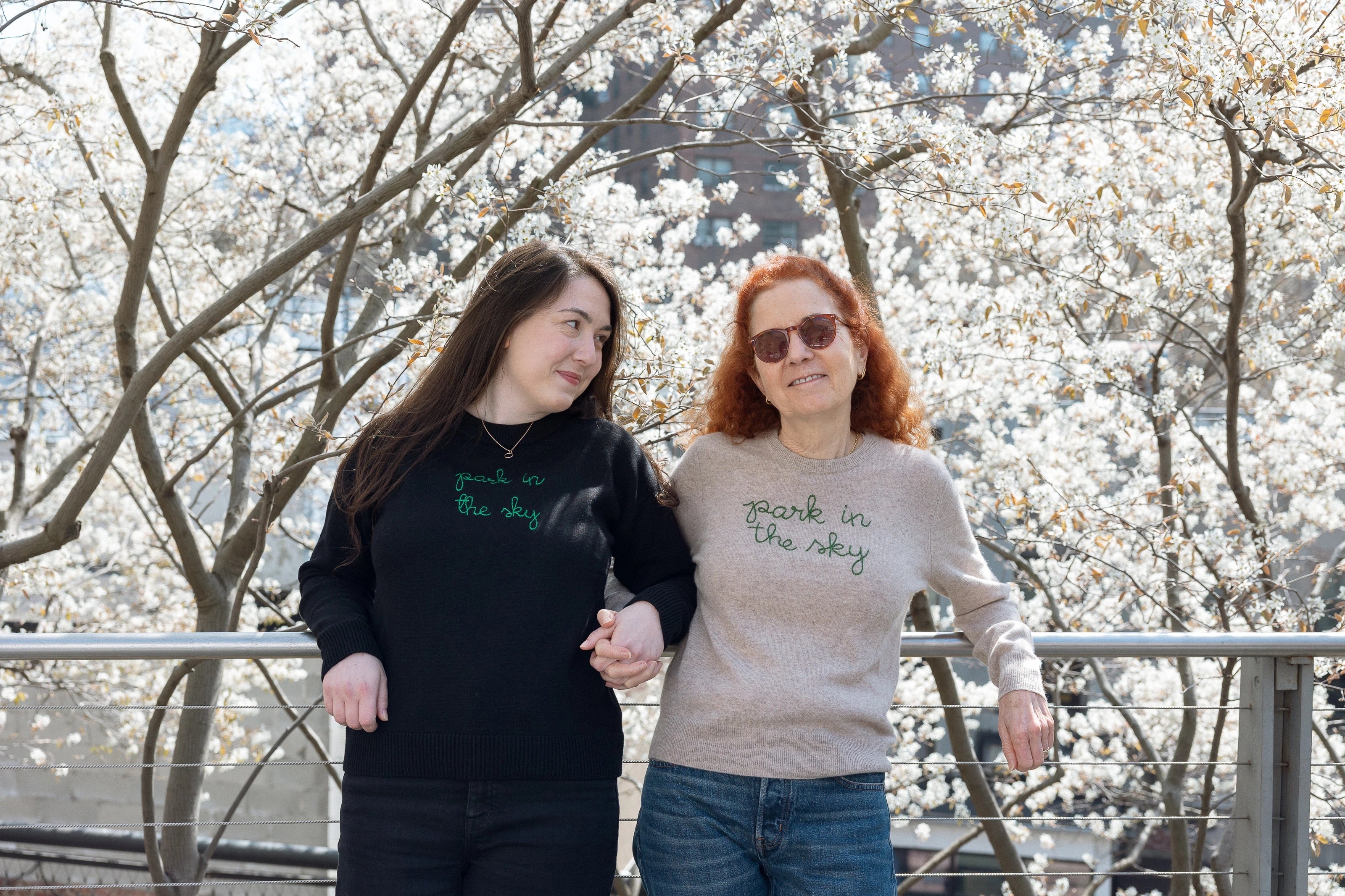 Mother and daughter stand amidst the serviceberry blooms on the High Line, holding hands. They're each wearing a Lingua Franca x High Line Sweater. The daughter is wearing the Maxine in Black and her mother is wearing the 100% Cashmere Sweater.