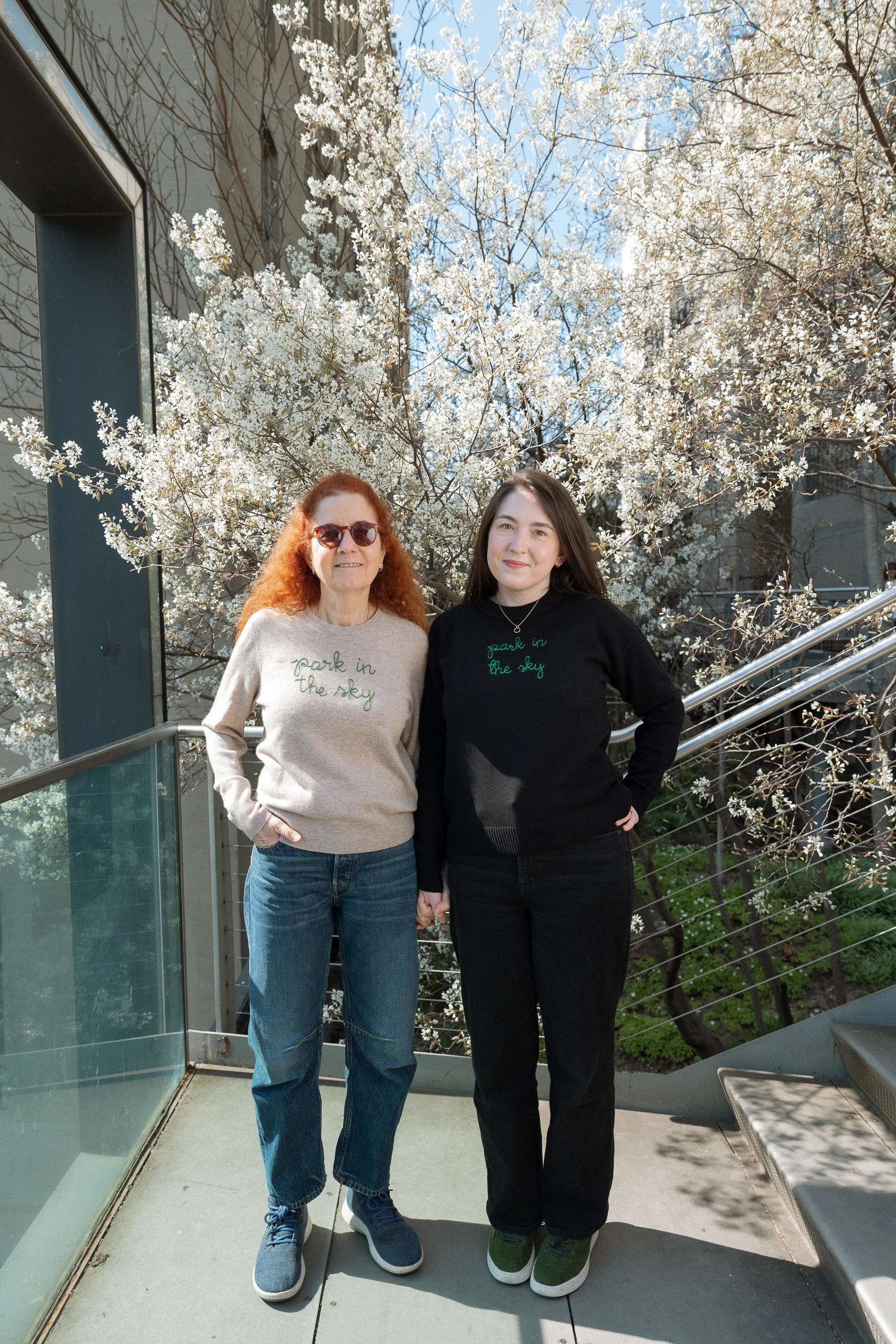 Mother and daughter standing at the 26th Street Overlook in front of the Serviceberry blossoms. Each wears a Lingua Franca x High Line Park in the Sky sweater. 