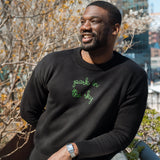 Man wears a Lingua Franca x High Line Maxine Sweater in Black, leaning on the railing of the High Line. This sweater is a cotton-cashmere blend, gender neutral and features the phrase "park in the sky" embroidered in green cursive on the chest. 