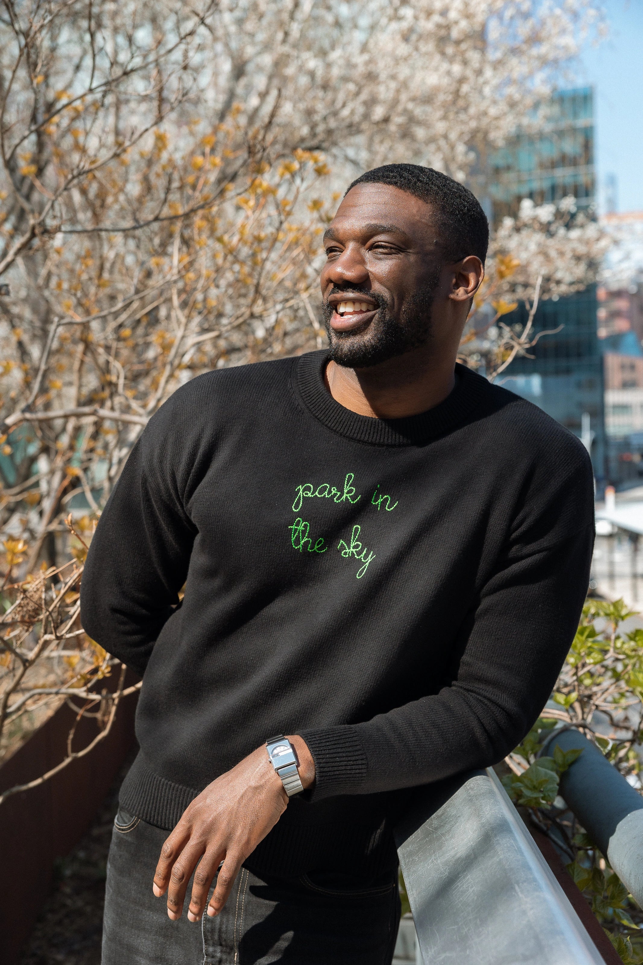Man wears a Lingua Franca x High Line Maxine Sweater in Black, leaning on the railing of the High Line. This sweater is a cotton-cashmere blend, gender neutral and features the phrase "park in the sky" embroidered in green cursive on the chest. 