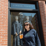 Woman wearing the Lingua Franca x High Line Maxine Sweater in navy. This cotton cashmere sweater is gender neutral and features the phrase "park in the sky" embroidered in green cursive on the chest. She is walking past the Lingua Franca store on Washington Street.