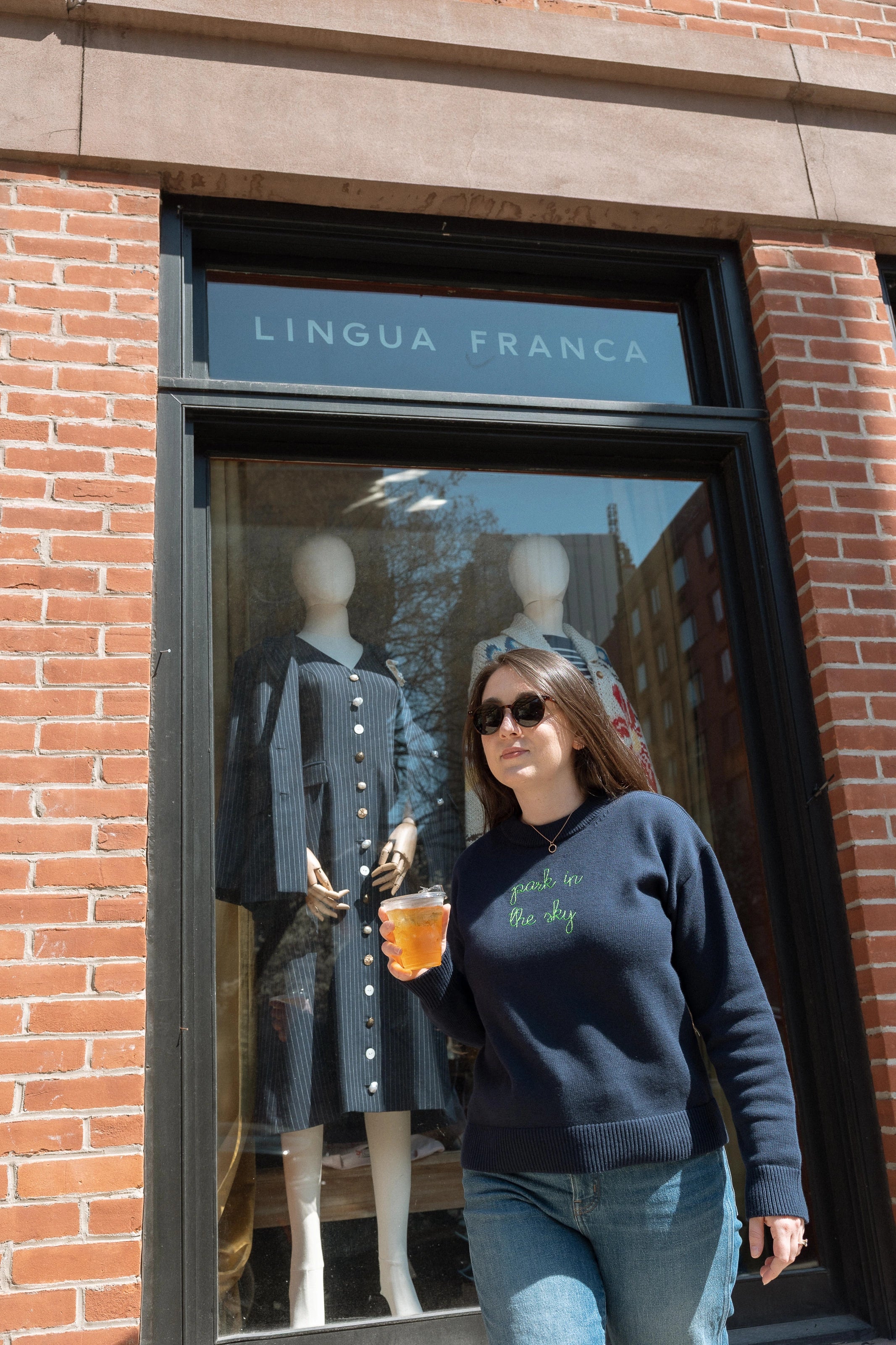 Woman wearing the Lingua Franca x High Line Maxine Sweater in navy. This cotton cashmere sweater is gender neutral and features the phrase "park in the sky" embroidered in green cursive on the chest. She is walking past the Lingua Franca store on Washington Street.
