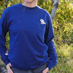 Person wearing the Pigeon Pocket Tee in Navy. A deep, saturated blue long-sleeve tee with a pocket on the left chest. An embroidered pigeon head peeks out of the pocket. On the right chest is a small embroidered High Line icon, a rail-road track inspired "H," in monochromatic thread. She stands on the High Line, foliage and rail both visible behind her.