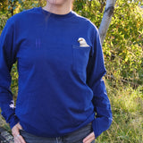 Person wearing the Pigeon Pocket Tee in Navy. A deep, saturated blue long-sleeve tee with a pocket on the left chest. An embroidered pigeon head peeks out of the pocket. On the right chest is a small embroidered High Line icon, a rail-road track inspired "H," in monochromatic thread. She stands on the High Line, foliage and rail both visible behind her.