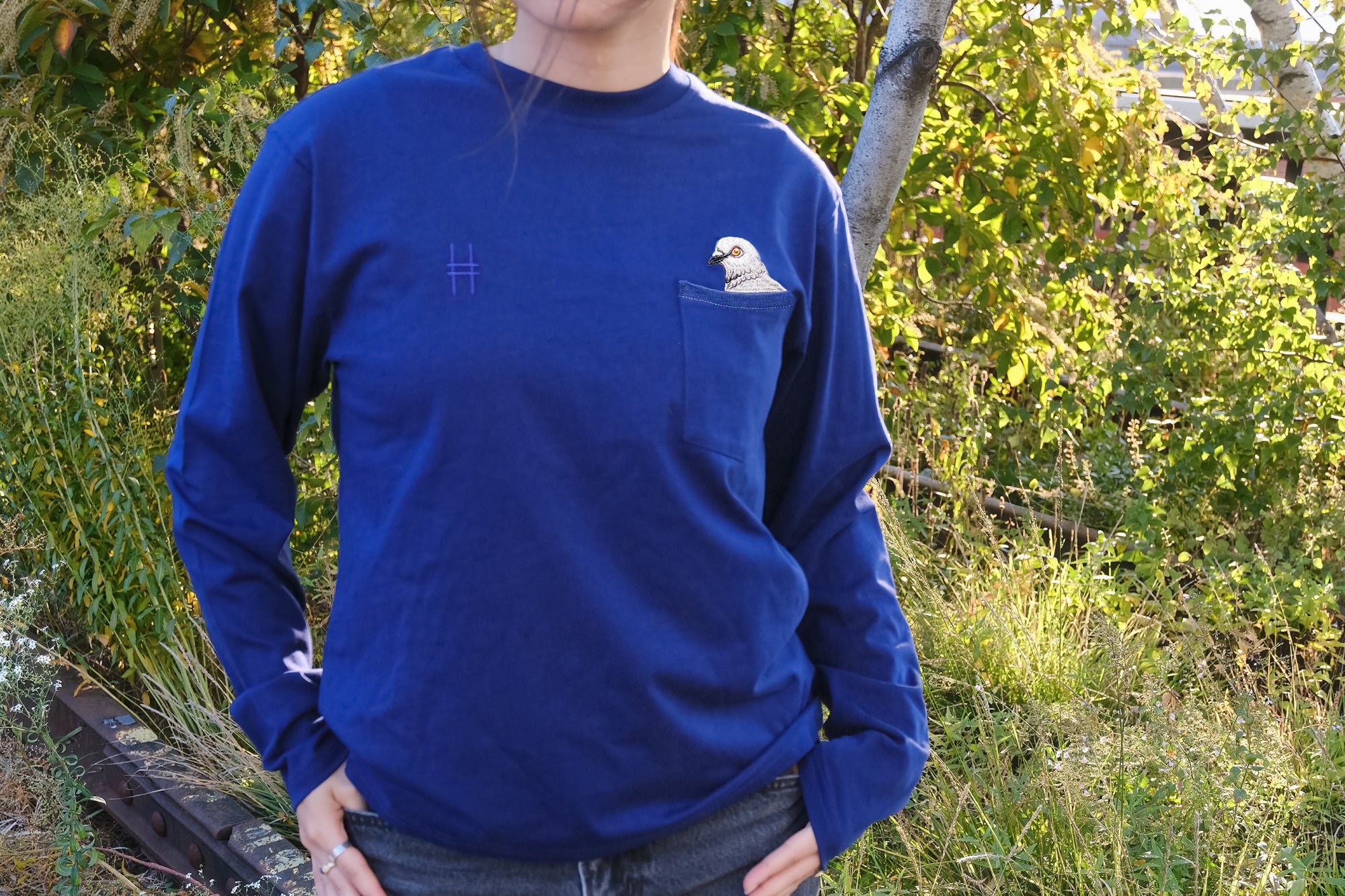 Person wearing the Pigeon Pocket Tee in Navy. A deep, saturated blue long-sleeve tee with a pocket on the left chest. An embroidered pigeon head peeks out of the pocket. On the right chest is a small embroidered High Line icon, a rail-road track inspired "H," in monochromatic thread. She stands on the High Line, foliage and rail both visible behind her.