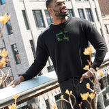 Man wears a Lingua Franca x High Line Maxine Sweater in Black, leaning on the railing of the High Line. This sweater is a cotton-cashmere blend, gender neutral and features the phrase "park in the sky" embroidered in green cursive on the chest.