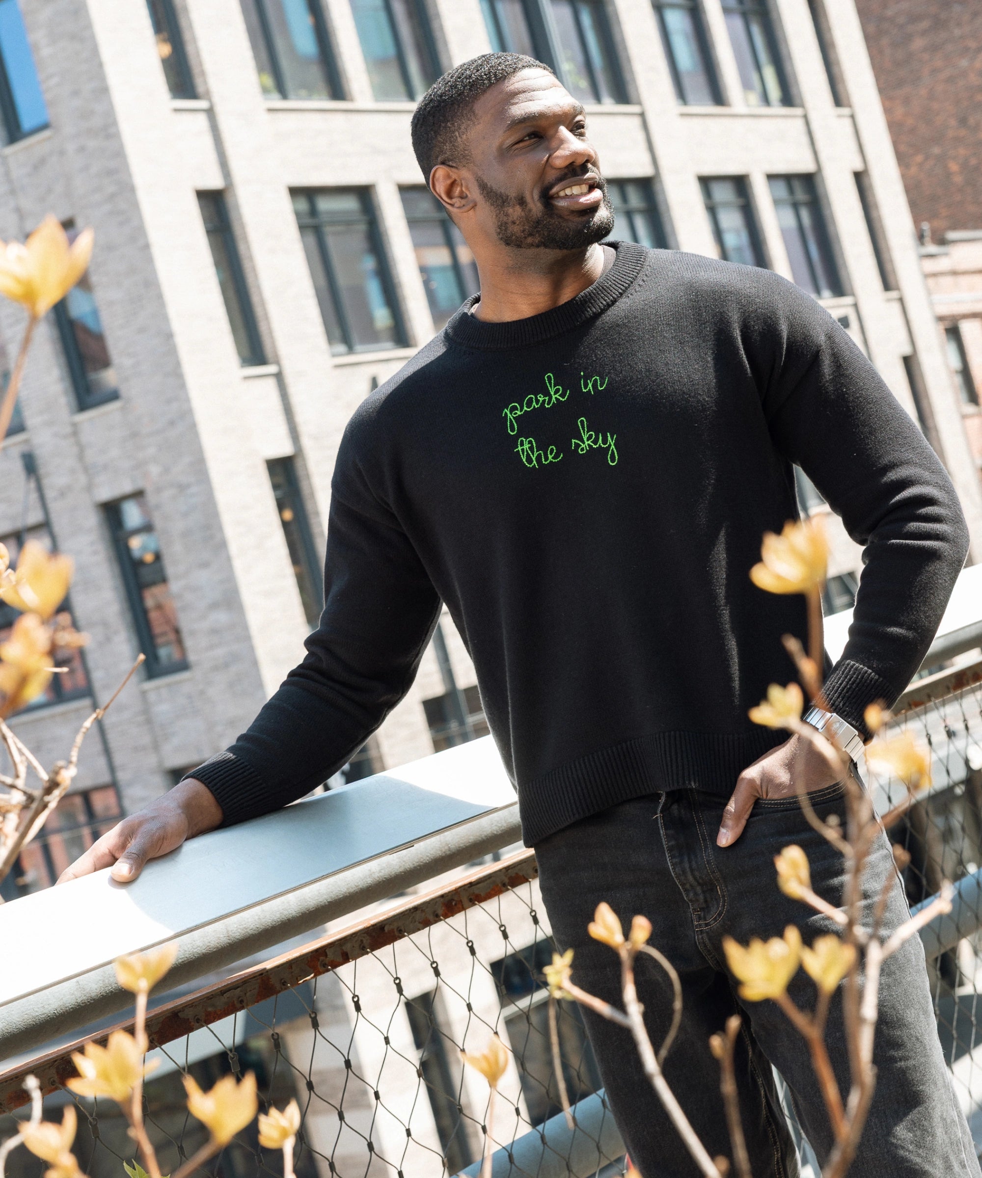 Man wears a Lingua Franca x High Line Maxine Sweater in Black, leaning on the railing of the High Line. This sweater is a cotton-cashmere blend, gender neutral and features the phrase "park in the sky" embroidered in green cursive on the chest.