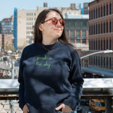 Woman stands on the Tiffany Overlook on the High Line and smiles while feeling the sun on her face through the trees. She wears the Lingua Franca x High Line Maxine Sweater in navy. This cotton cashmere sweater is gender neutral and features the phrase "park in the sky" embroidered in green cursive on the chest.