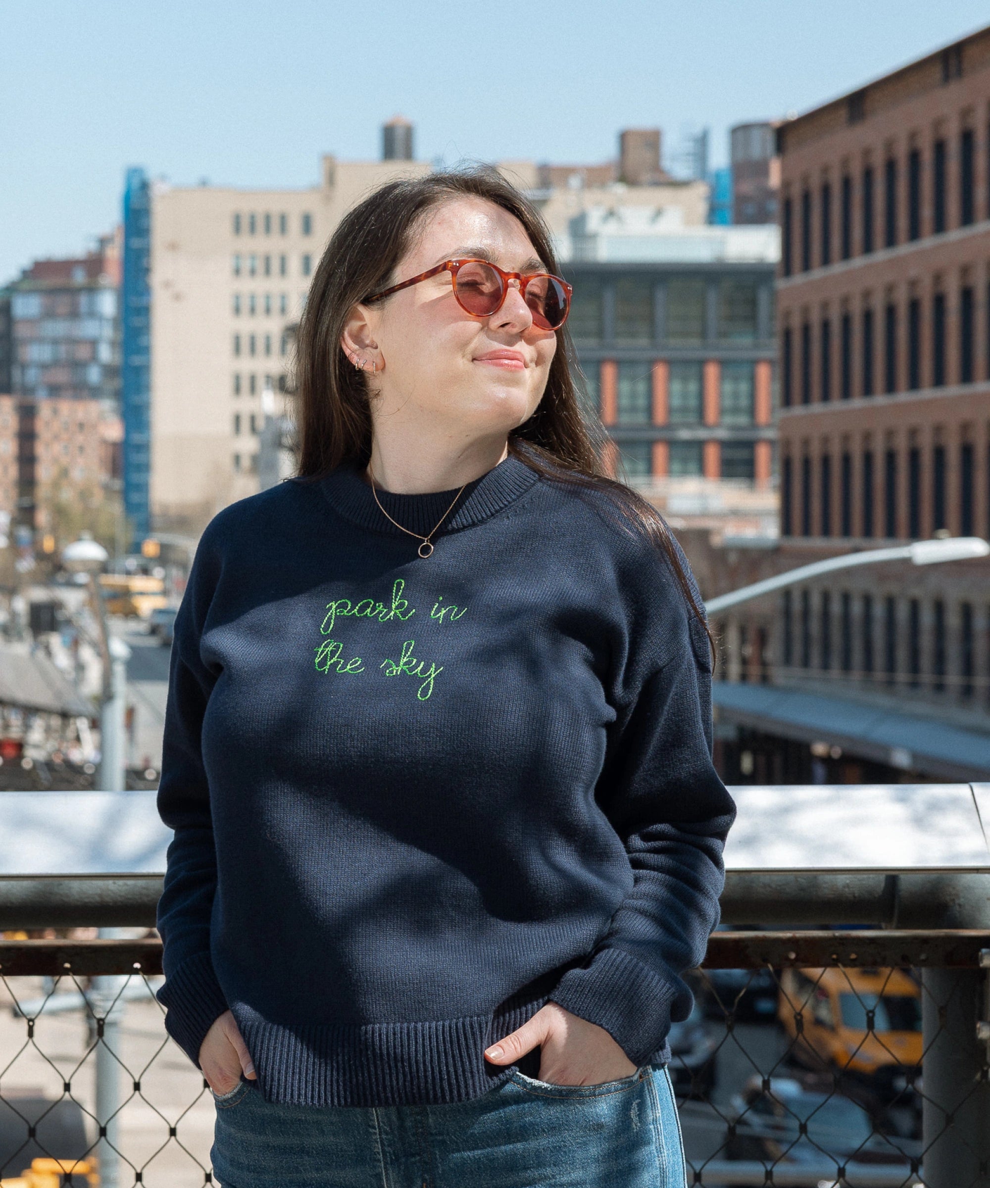 Woman stands on the Tiffany Overlook on the High Line and smiles while feeling the sun on her face through the trees. She wears the Lingua Franca x High Line Maxine Sweater in navy. This cotton cashmere sweater is gender neutral and features the phrase "park in the sky" embroidered in green cursive on the chest.