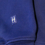 Closeup on the High Line icon, a railroad track inspired "H" on a small woven navy patch sewn onto the sleeve of the Park in the Sky Crewneck.