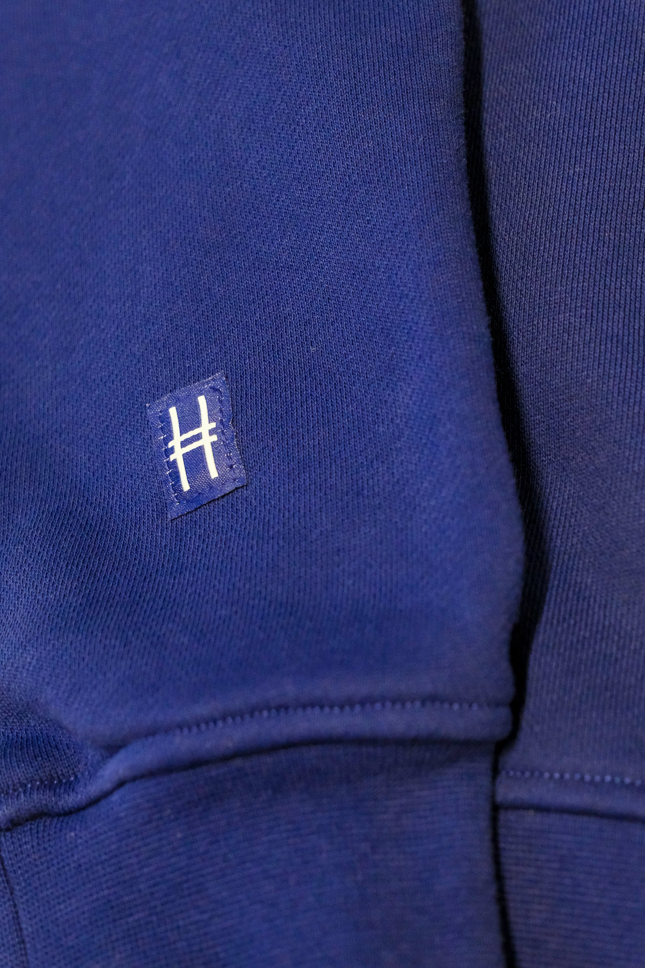 Closeup on the High Line icon, a railroad track inspired "H" on a small woven navy patch sewn onto the sleeve of the Park in the Sky Crewneck.