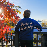 Person stands, leaning on the rail of the High Line at 14th street looking out at the Hudson River.  The sun shines through vibrant red and orange sumac tree to their left. They wear the Park in the Sky Crewneck in Navy, a french terry cotton embroidered crewneck sweatshirt with "Park in the Sky" in script across the back.