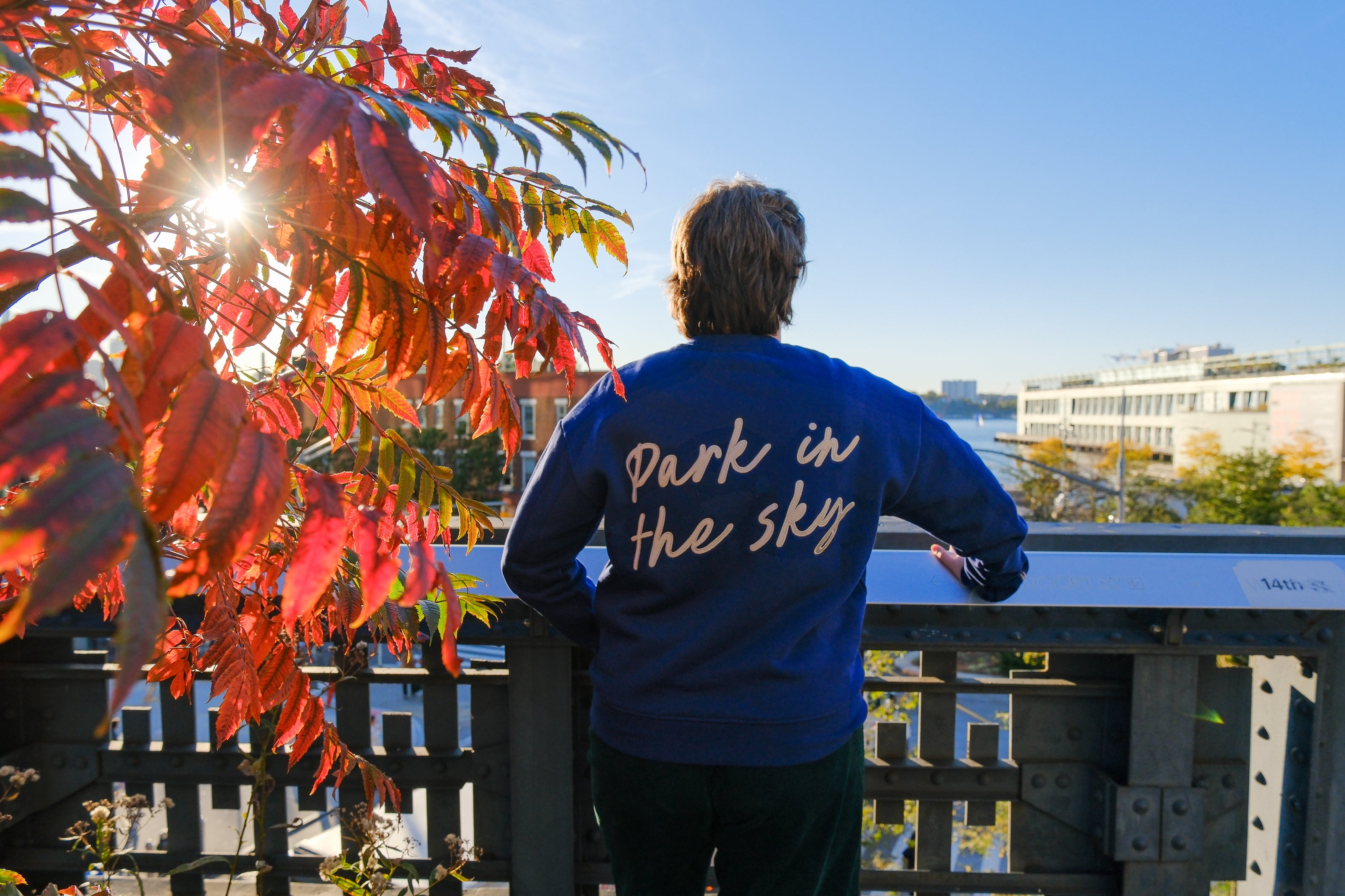 Person stands, leaning on the rail of the High Line at 14th street looking out at the Hudson River.  The sun shines through vibrant red and orange sumac tree to their left. They wear the Park in the Sky Crewneck in Navy, a french terry cotton embroidered crewneck sweatshirt with "Park in the Sky" in script across the back.