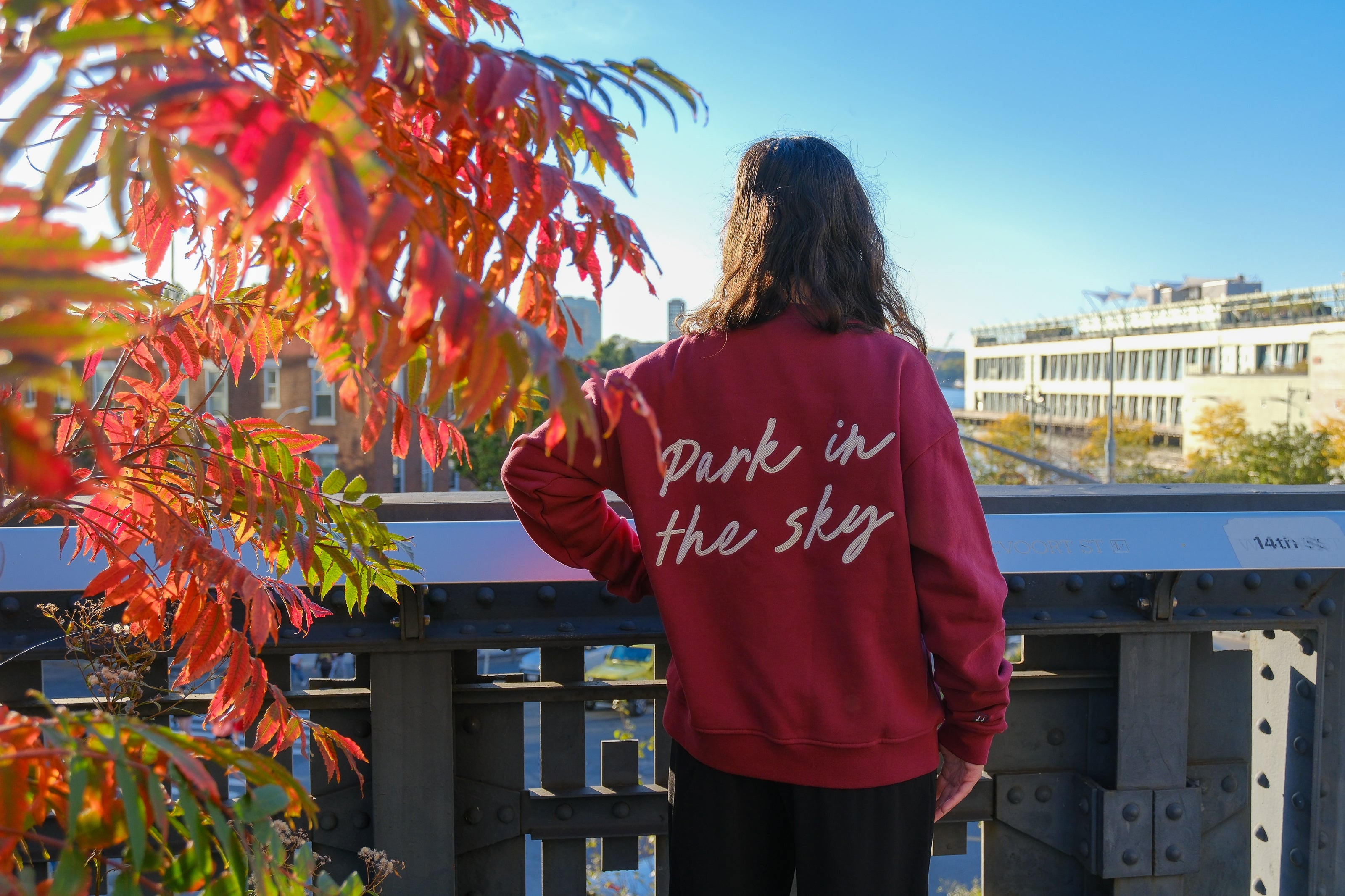 Woman stands, left hand on her hip, on the High Line at 14th street looking out at the Hudson River, a vibrant red and orange sumac tree to her left. She wears the Park in the Sky Crewneck in Smokebush, a french terry cotton embroidered crewneck sweatshirt in a deep red with "Park in the Sky" in script in white across the back.