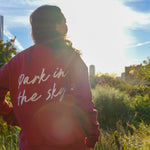 Woman stands, left hand on her hip, on the High Line looking at the lush foliage, the Whitney Museum in the background. She wears the Park in the Sky Crewneck in Smokebush, a french terry cotton embroidered crewneck sweatshirt in a deep red with "Park in the Sky" in script in white across the back.