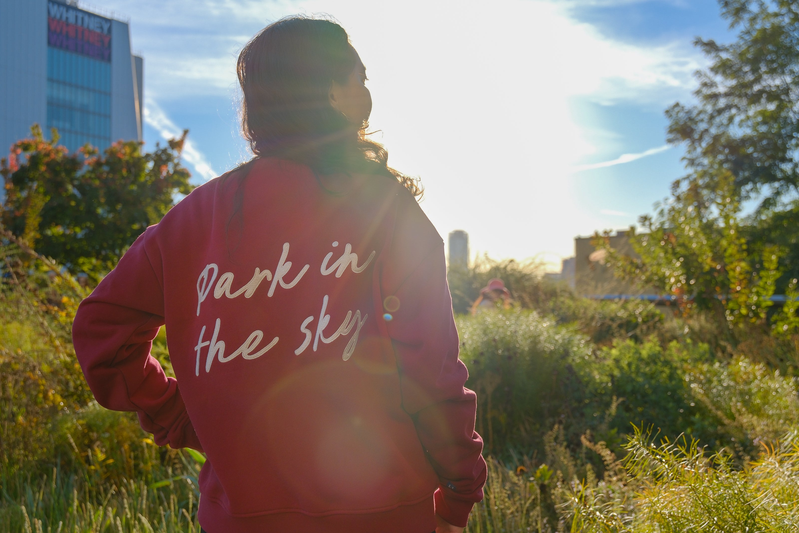 Woman stands, left hand on her hip, on the High Line looking at the lush foliage, the Whitney Museum in the background. She wears the Park in the Sky Crewneck in Smokebush, a french terry cotton embroidered crewneck sweatshirt in a deep red with "Park in the Sky" in script in white across the back.