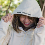 Woman wearing Park in the Sky hoodie, pulling the hood up so the "Park in the Sky" embroidery detail on the hood is visible.
