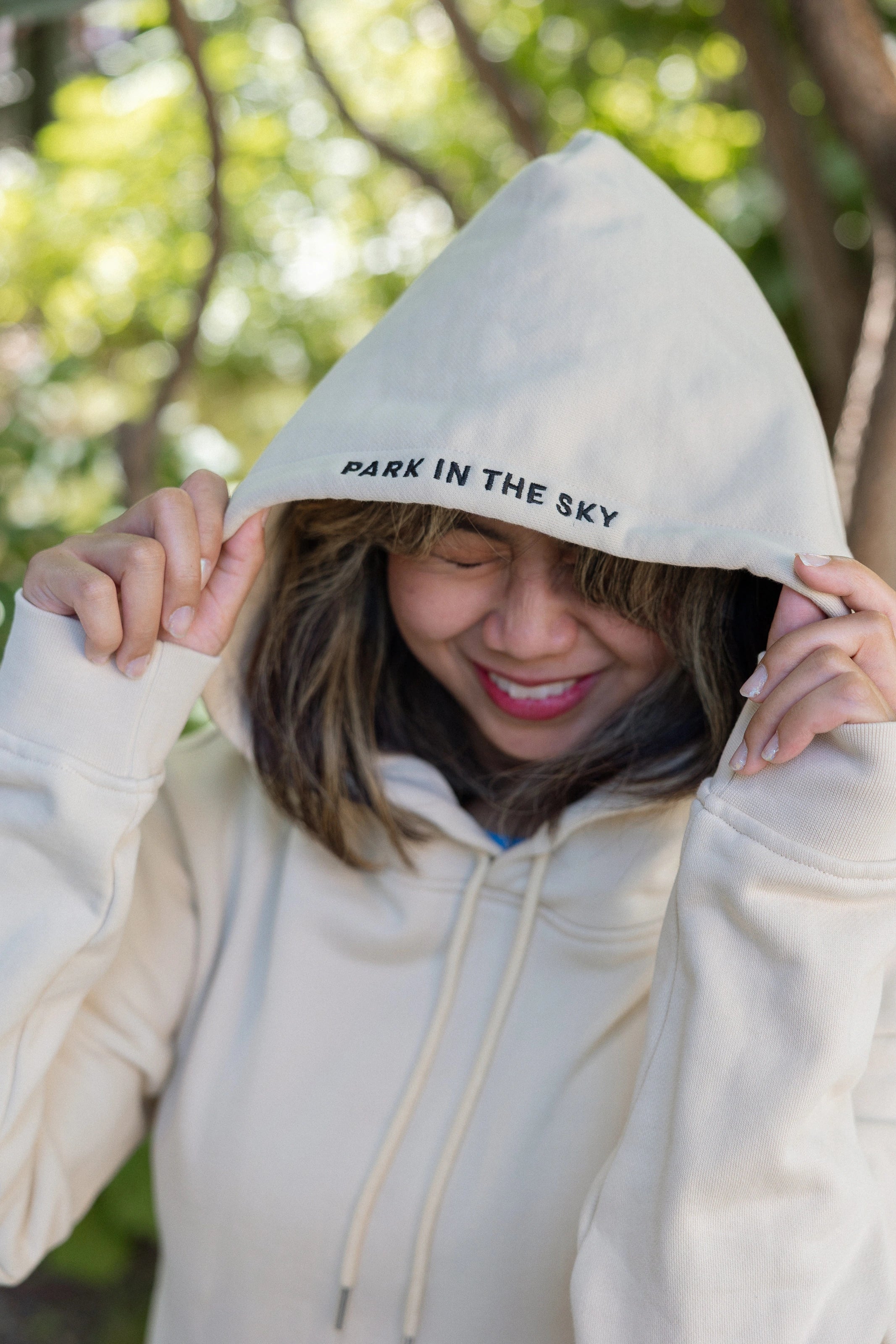 Woman wearing Park in the Sky hoodie, pulling the hood up so the "Park in the Sky" embroidery detail on the hood is visible.