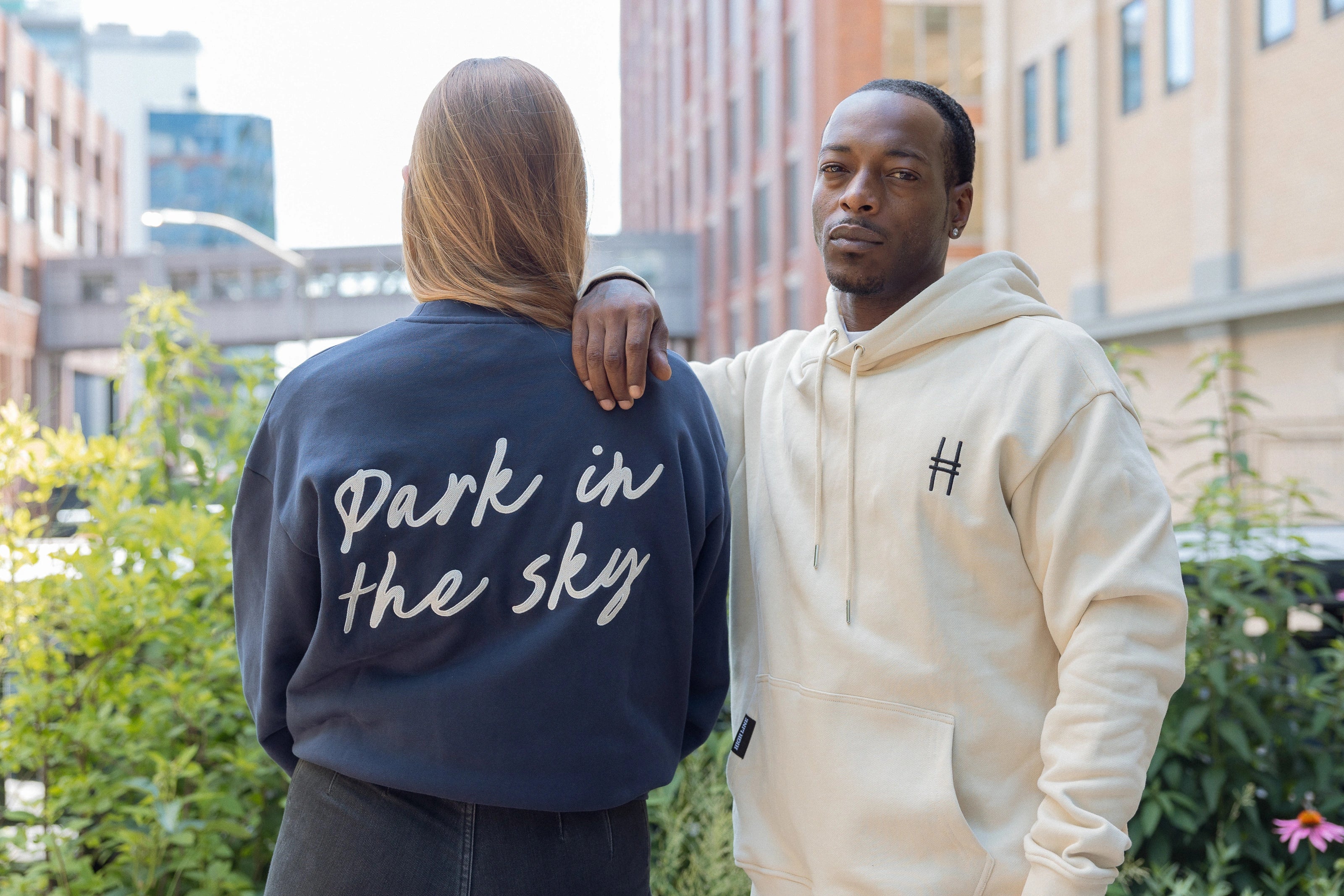 Woman wearing Park in the Sky crewneck and man in the Park in the Sky hoodie, with crewneck back details and front icon embroidery on the hoodie visible.