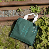 Park in the Sky Tote in Ivy with all the Community in the Sky collection pins on it, resting against the railroad tracks of the High Line.