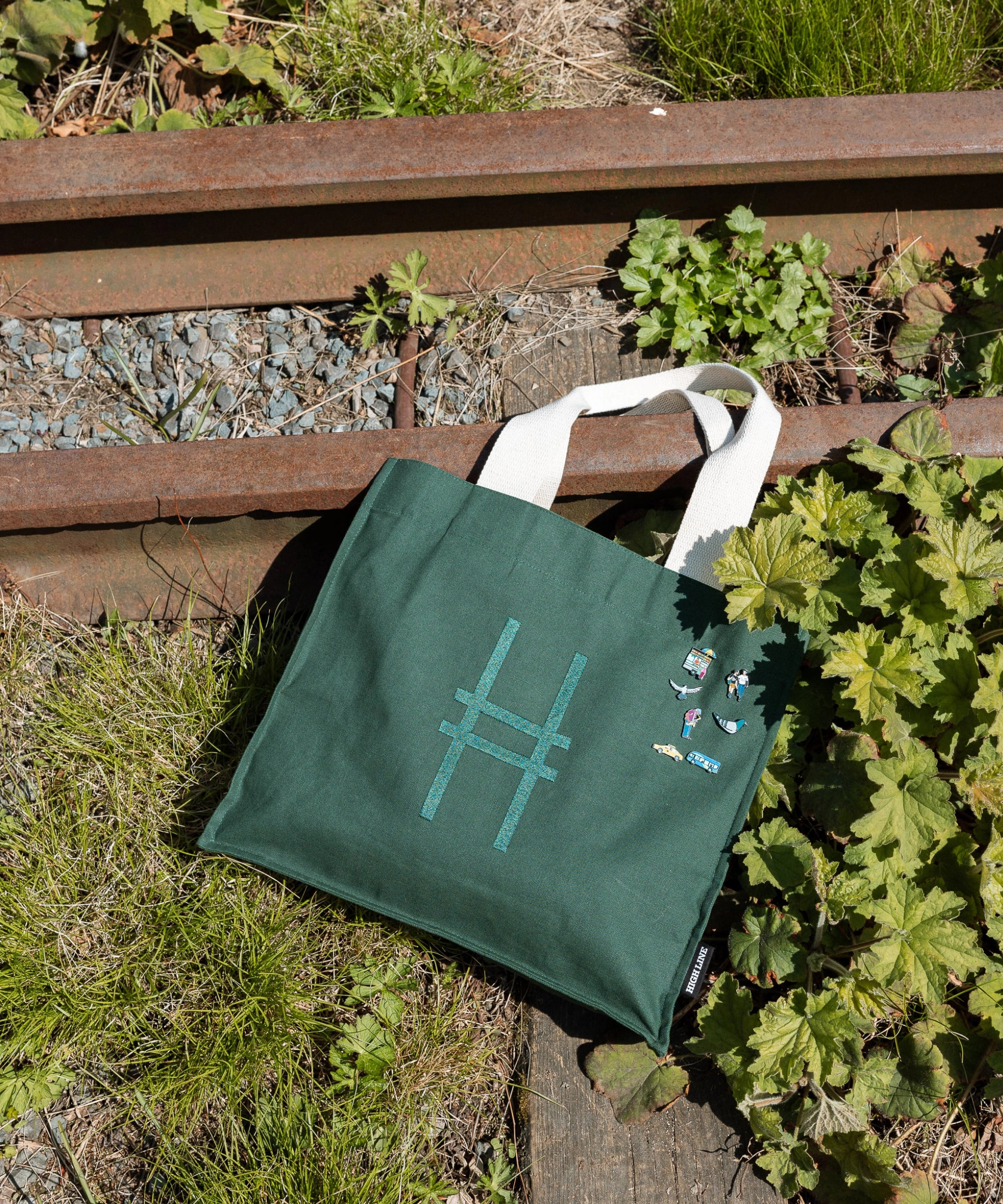 Park in the Sky Tote in Ivy with all the Community in the Sky collection pins on it, resting against the railroad tracks of the High Line.
