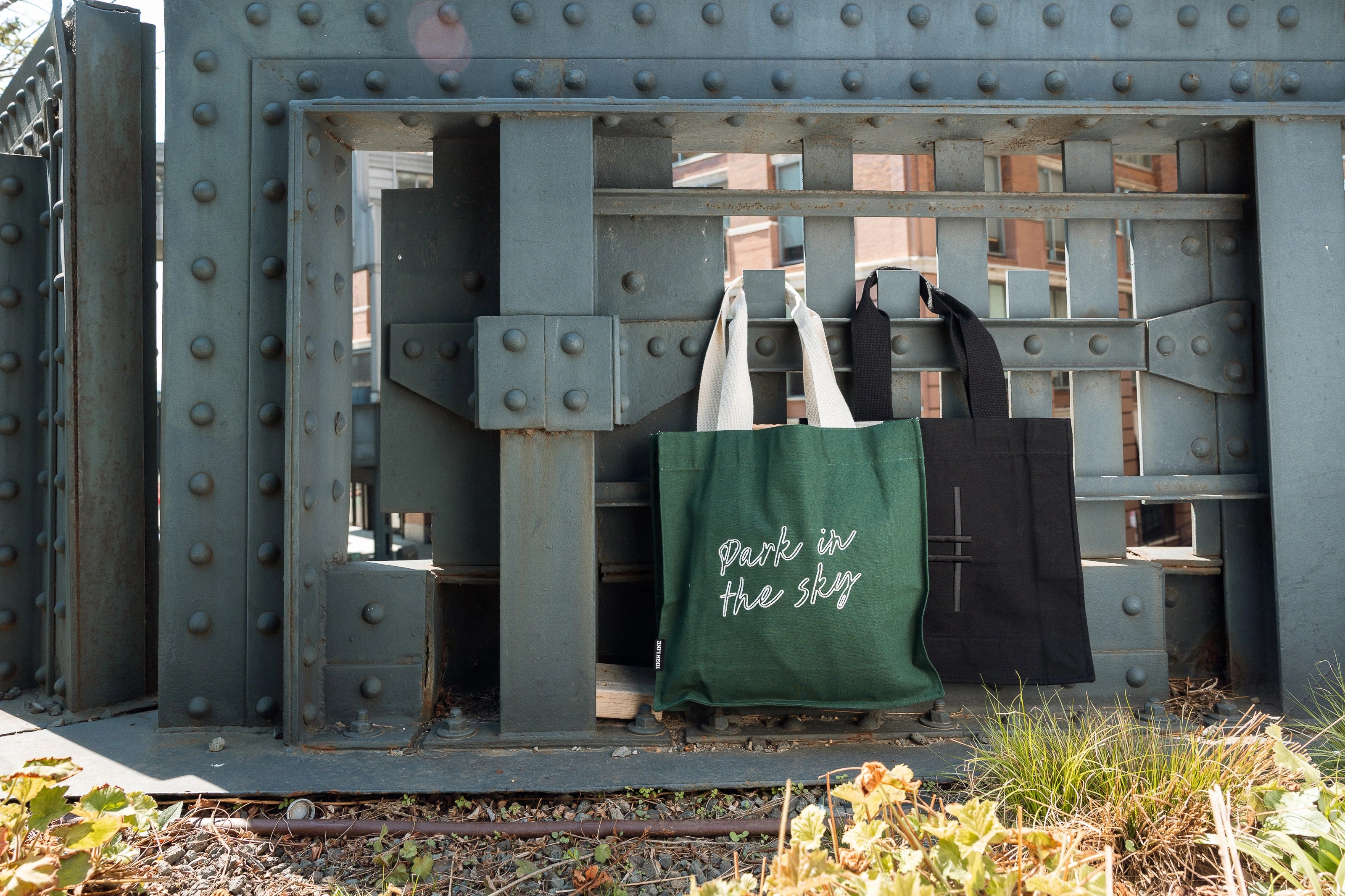 Park in the Sky Tote in Ivy and Park in the Sky Tote in Black hanging off of the railing of the High Line.