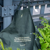 Piet Oudolf Quote Apron in dark green, featuring the High Line icon and the quote "The garden is a promise. And I think that promise is why you want to come back." in a pale yellow. The apron hangs off of the High Line rail and is nestled amongst plants.