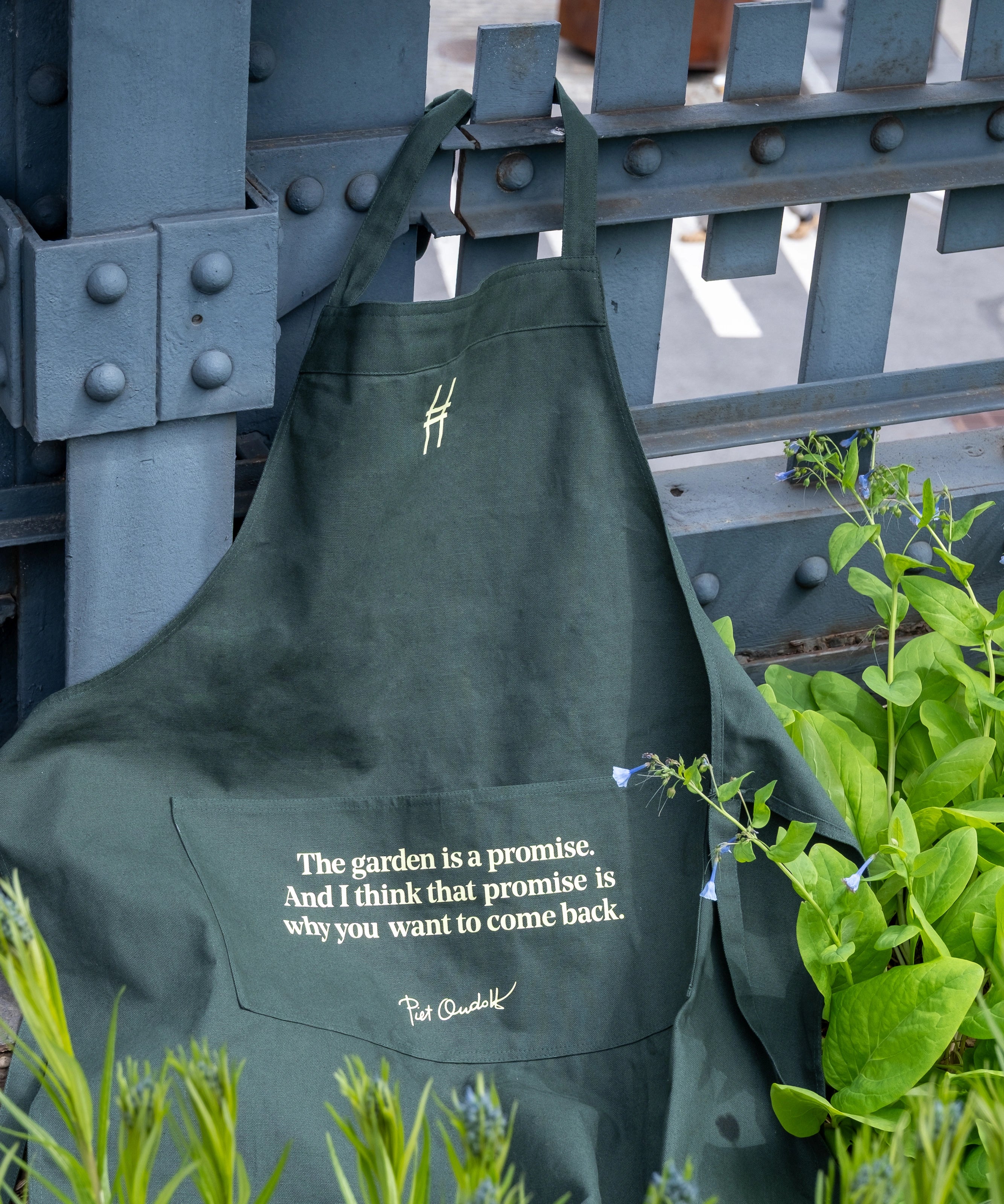 Piet Oudolf Quote Apron in dark green, featuring the High Line icon and the quote "The garden is a promise. And I think that promise is why you want to come back." in a pale yellow. The apron hangs off of the High Line rail and is nestled amongst plants.