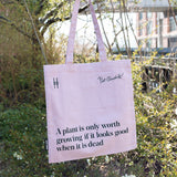 Piet Oudolf Quote Tote in Aster—a pale purple cotton canvas tote bag featuring the quote "A plant is only worth growing if it looks good when it is dead." The tote is being held up against the foliage of the High Line.